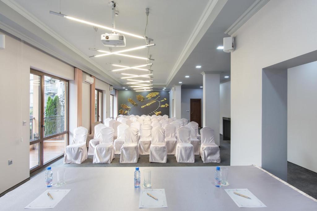 Konferenču telpa ar sēdvietām, kas klātas ar baltām segām, un modernu apgaismojumu. Redzams projekcijas ekrāns un galds ar rakstāmpiederumiem.