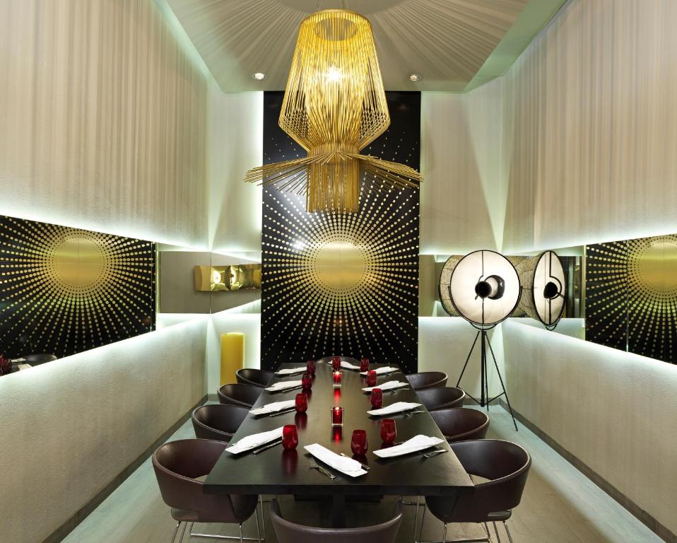 Mūsdienīgs restorāns ar elegantu interjeru, kurā ir liels galds, apgaismots ar stilīgu lampu un atspoguļojumiem uz sienām. Sarkanās sveces papildina atmosfēru.