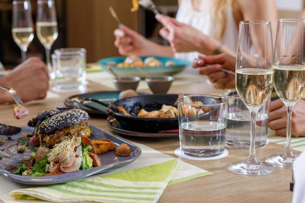 Eleganta maltīte ar dažādiem ēdieniem, tostarp melnā maizē gatavots burgeris un garnējumi, kopā ar šampanieti un ūdeni. Galda estētika ir mūsdienīga un pievilcīga.