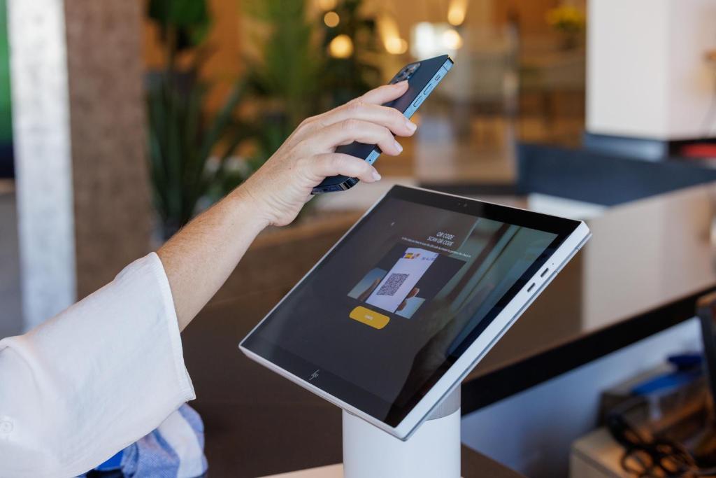 Rokas ar mobilo tālruni skenē QR kodu uz interaktīvas ierīces, kas atrodas reģistrācijas galdiņā. Moderns risinājums digitālajām mijiedarbībām.