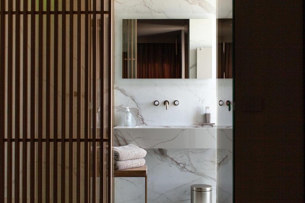 Mūsdienīgs vannas istabas interjers ar marmora sienām, spoguli un elegantu apgaismojumu. Redzamas dvieļu kaudze un higiēnas līdzekļi uz izlietnes.