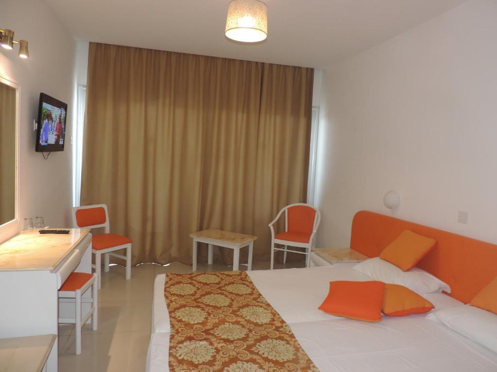 Gaiša viesnīcas numura iekārtojums ar oranžiem akcentiem, gultu un televizoru. Numurā ir arī galds un krēsli, kā arī dzeltenas aizkari.