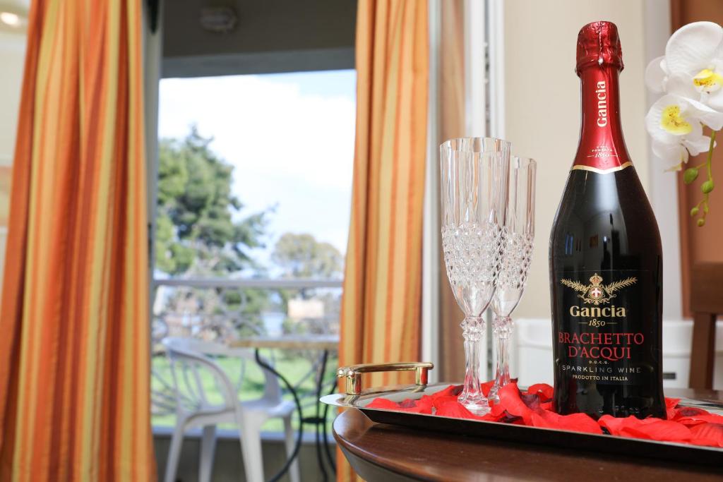 Šampanieša pudele "Gancia Brachetto d'Acqui" un divi glāzes uz galda, rotātas ar rožu ziedlapiņām, ar skatu uz dārzu caur atvērtajiem logiem.