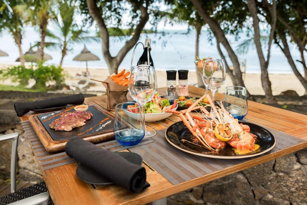 Elegantā pludmales restorāna galda kompozīcija ar gaļas un jūras velšu ēdieniem, dzērieniem un svaigiem dārzeņiem, fonā redzami palmas un pludmale.