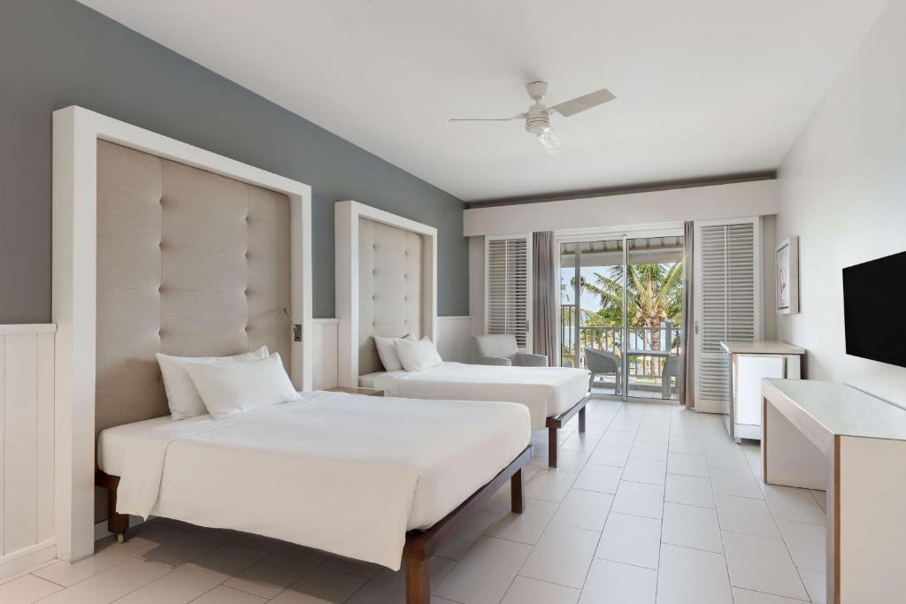 Mūsdienīgs viesnīcas numurs ar divām gultām, gaišām sienām un logiem, kas ved uz terasi ar palmām. Numurā ir televizors un moderns interjers.