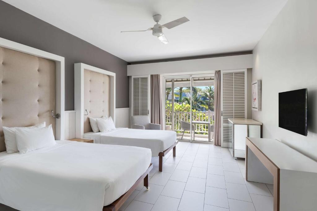 Mūsdienīgs viesnīcas numurs ar divām gultām, pelēkām sienām un lieliem logiem, kas ved uz balkonu ar skatu uz dārzu. Interjers ir gaišs un elegants, ar minimālistisku dizainu.