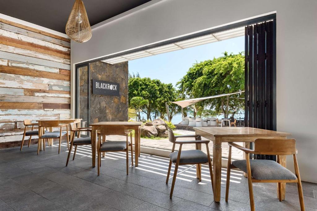 Mūsdienīgs restorāna interjers ar koka galdiem un krēsliem, kas piedāvā skatu uz zaļojošu dabu un pludmali. Gaismas caurlaidība un plašas durvis rada viesmīlīgu atmosfēru.