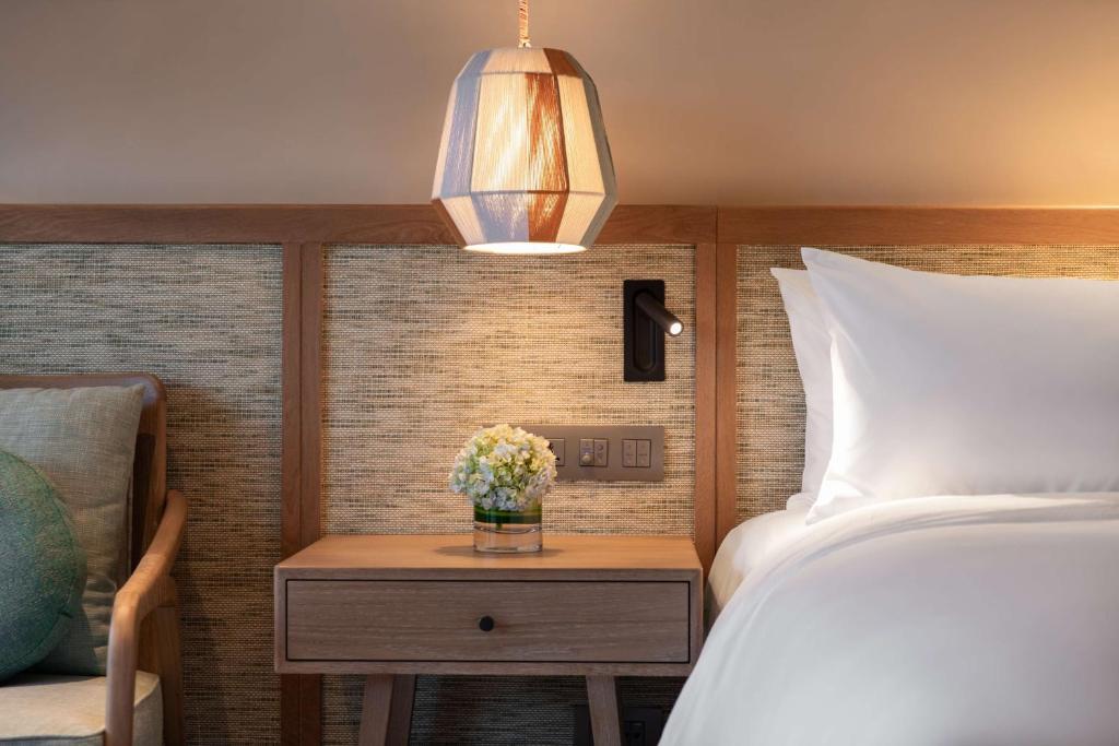 Mūsdienīgs guļamistabas interjers ar koka naktsgaldiņu, stilīgu lampu un ziedu kompozīciju. Svaigas krāsas un ērts dizains rada relaksējošu atmosfēru.