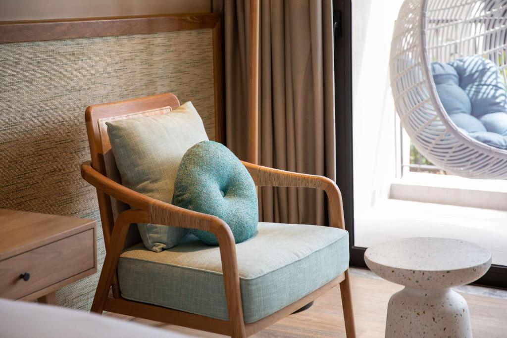 Mūsdienīgs viesnīcas numura stūris ar ērtu koka krēslu, kas rotāts ar zilu spilvenu un pievienotu mazu galdiņu. Loga tuvumā redzama šūpuļkrēsla un dabiska apgaismojuma atmosfēra.