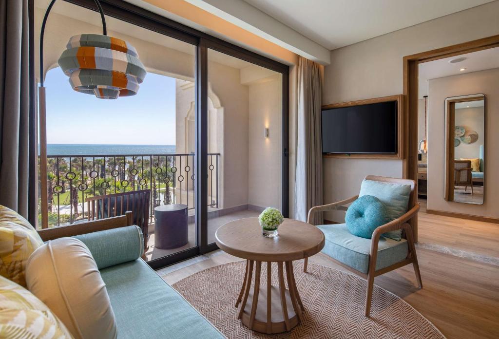Mūsdienīga viesnīcas istaba ar plašu skatu uz jūru, gaišām krāsām un ērtu mēbelējumu, tostarp dīvānu un atzveltnes krēslu. 