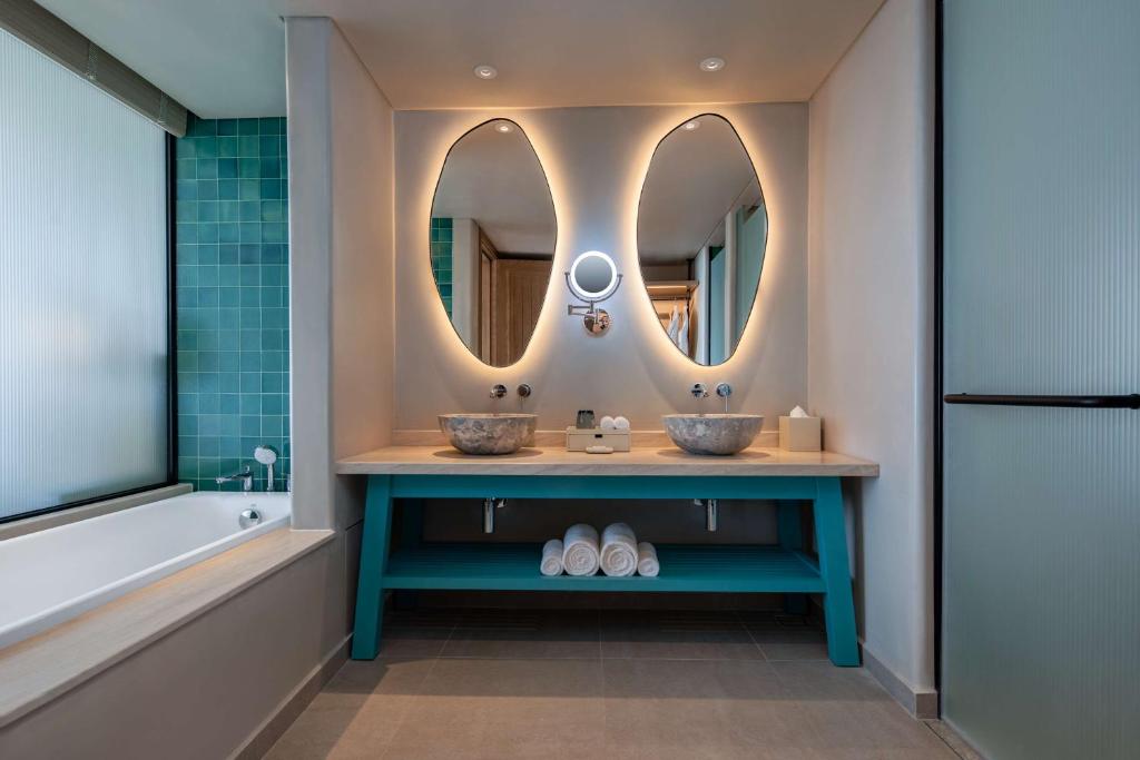 Mūsdienīgs vannas istabas interjers ar diviem ovāliem spoguļiem, akmens izlietni un tirkīza krāsas galdu. Attēlā redzama arī vanna un stikla durvis.