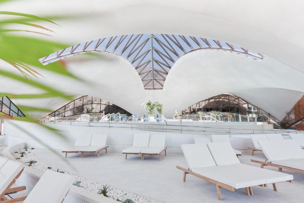 Mūsdienīgs un elegants spa interjers ar baltiem sauļošanās krēsliem un lieliem logiem, kas nodrošina daudz gaismas. Telpa ir veidota ar mūsdienīgu arhitektūru, radot relaksējošu atmosfēru.