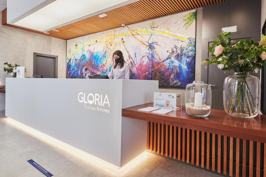 Reģistratūra viesnīcā "Gloria Thalasso & Hotels" ar modernu dizainu un krāsainu mākslas darbu uz sienas. Uz galda ir ziedu kompozīcija un informatīvie materiāli.