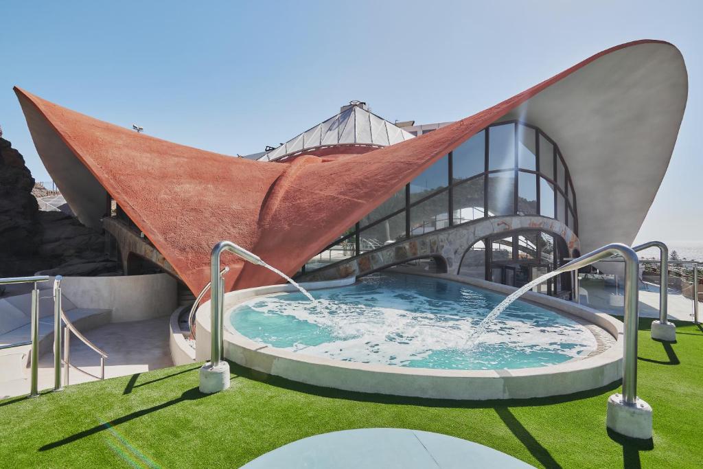 Mūsdienīga arhitektūra ar viļņveida jumtu un apaļu baseinu, kas atrodas pie jūras. Dizains apvieno dabas elementus ar inovatīvām konstrukcijām.