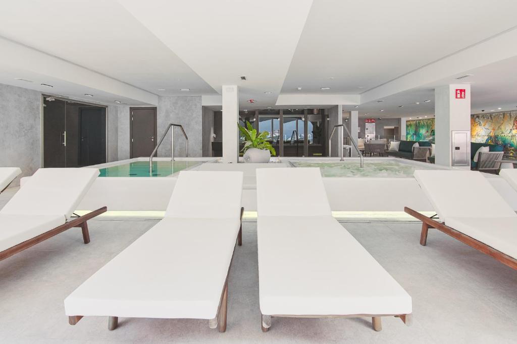 Mūsdienīgs spa telpas interjers ar baltām sauļošanās krēslām un relaksējošu baseinu. Pēcpusdienas gaisma un zaļumi rada mierīgu atmosfēru.