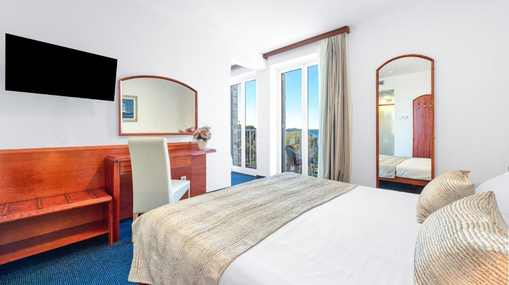 Moderns viesnīcas numurs ar gultu, galdu, krēslu un televizoru pie sienas. Lielas logu durvis nodrošina dabīgu apgaismojumu un skatu uz apkārtni.