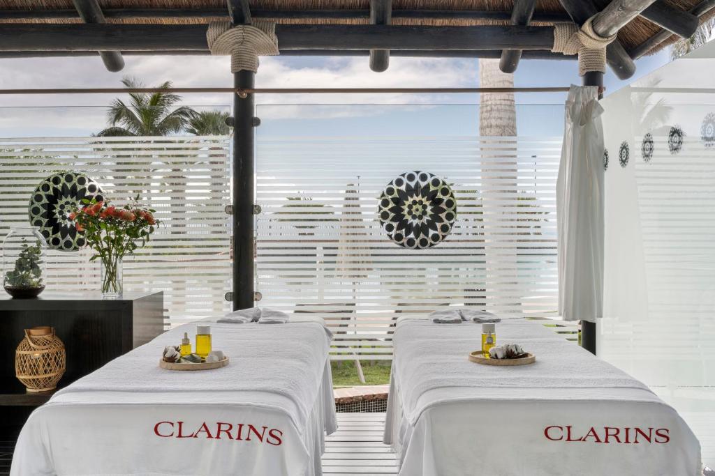 Relaksējoša spa telpa ar diviem masāžas galdiem, dekorētiem ar uzņēmuma "Clarins" logo, un dabīgām ainavām fonā. Uz galdiem atrodas masāžas eļļas un rotājumi.