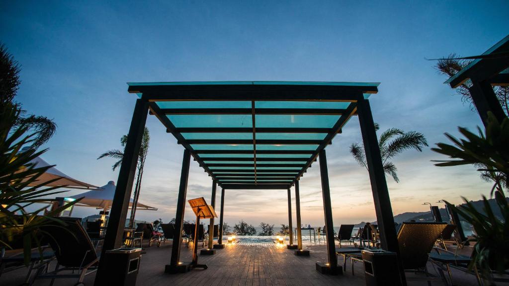 Mūsdienīga terase ar stikla jumtu, no kuras paveras skats uz saulrietu pie peldbaseina. Apkārt ir palmām un atpūtas krēsliem, radot relaksējošu atmosfēru.