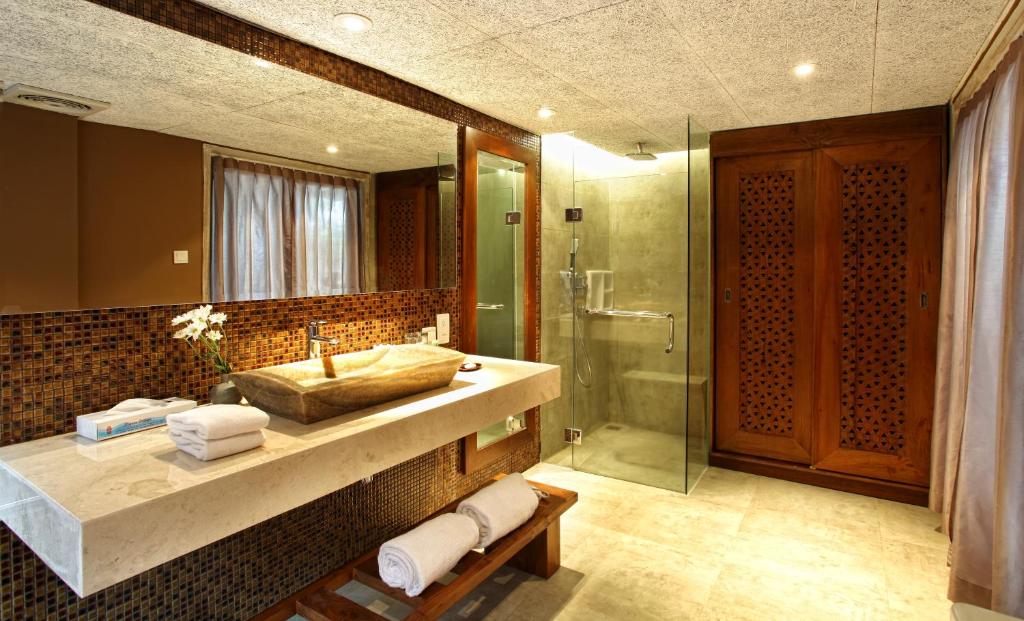 Mūsdienīgs vannas istabas interjers ar akmens izlietni, stikla dušu un koka detalēm, radot relaksējošu atmosfēru. Telpa ir gaiša un eleganta, ar flīžu apdari un ziediem uz izlietnes.