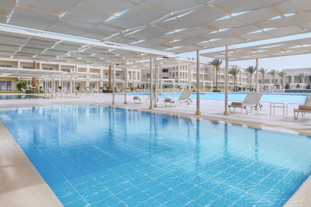 Lieliska baseina zona ar saulessargiem un atpūtas krēsliem, kas atrodas pie viesnīcas kompleksa. Skats uz dzidru ūdeni un palmām, radot relaksējošu atmosfēru.