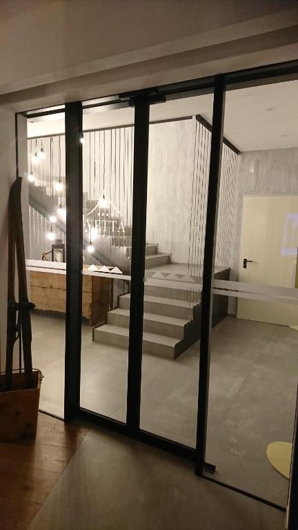 Mūsdienīgs interjers ar stikla durvīm, kas ved uz gaišu kāpņu telpu, apgaismotu ar stilīgām lampām. Redzama dekoratīva siena un koka mēbeles.