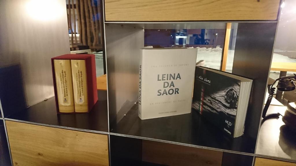 Grāmatu plaukts ar trim grāmatām, tostarp "Leina da Saor" un citiem izdevumiem, kā arī klasiskā stila telefonu.