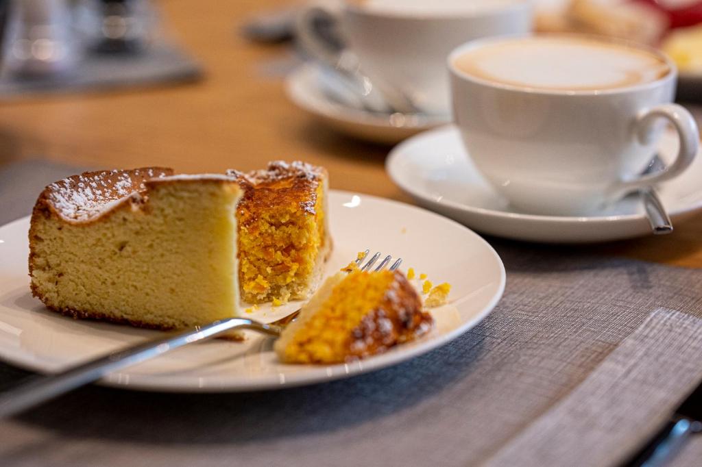 Saldais kūkas gabals, kas sagriezts un pasniegts uz baltas šķīvja blakus tasei ar kafiju. Galds ir klāts ar pelēku audumu, radot viesmīlīgu atmosfēru.