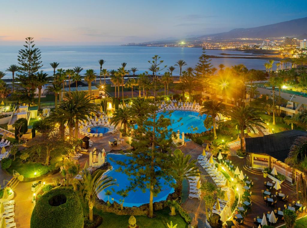 Tropu viesnīca ar baseiniem un palmām, kas atrodas pie jūras, izgaismota saulrieta gaismā. Skats uz okeānu un pilsētas apgaismojumu fonā.