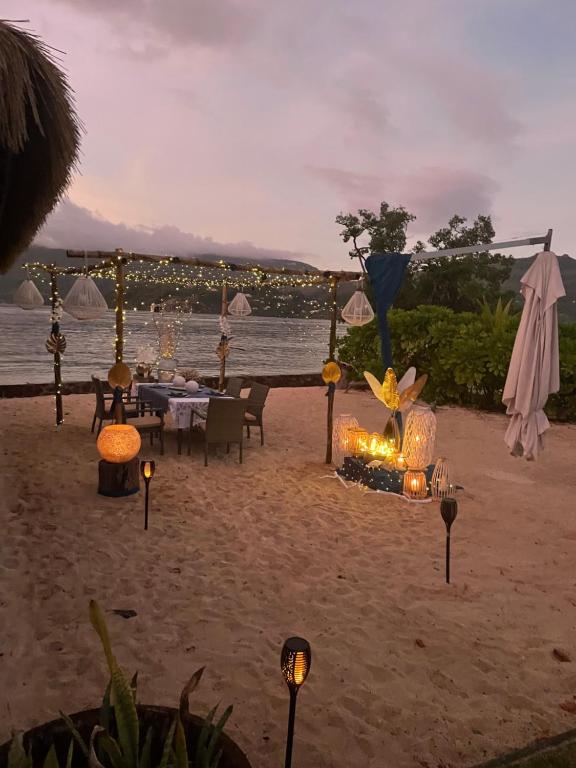 Pludmales restorāna romantiskais galdiņš, apgaismots ar svecēm un dekoratīvām lampām, pretī jūrai un kalniem saulrieta laikā.