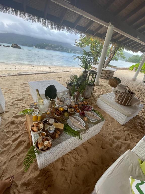 Pludmales restorāna galda klāšana ar dažādiem gardumiem, dzērieniem un dekoratīviem elementiem, fons ar jūru un kalniem.