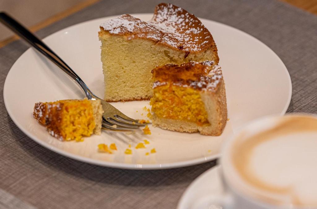 Gards kūka gabals ar dzeltenu pildījumu, apkaisīts ar pūdercukuru, pasniegts uz baltas šķīvja blakus dakšiņai un kafijai.