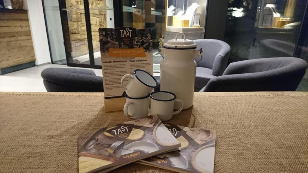 Kafijas galda noformējums ar tradicionālajiem krūzēm, balto piena trauku un ēdienkarti. Aiz fonā redzami mūsdienīgi krēsli un koka sienas elementi.