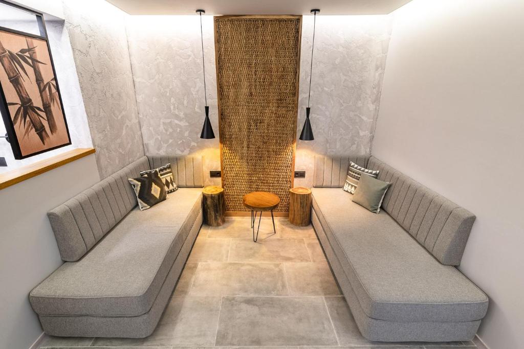 Mūsdienīgs interjers ar divām pelēkām dīvānu zonām, koka sēdekli un siltu apgaismojumu, radot relaksējošu atmosfēru. Sienā ir mākslas darbs un teksturēta virsma.