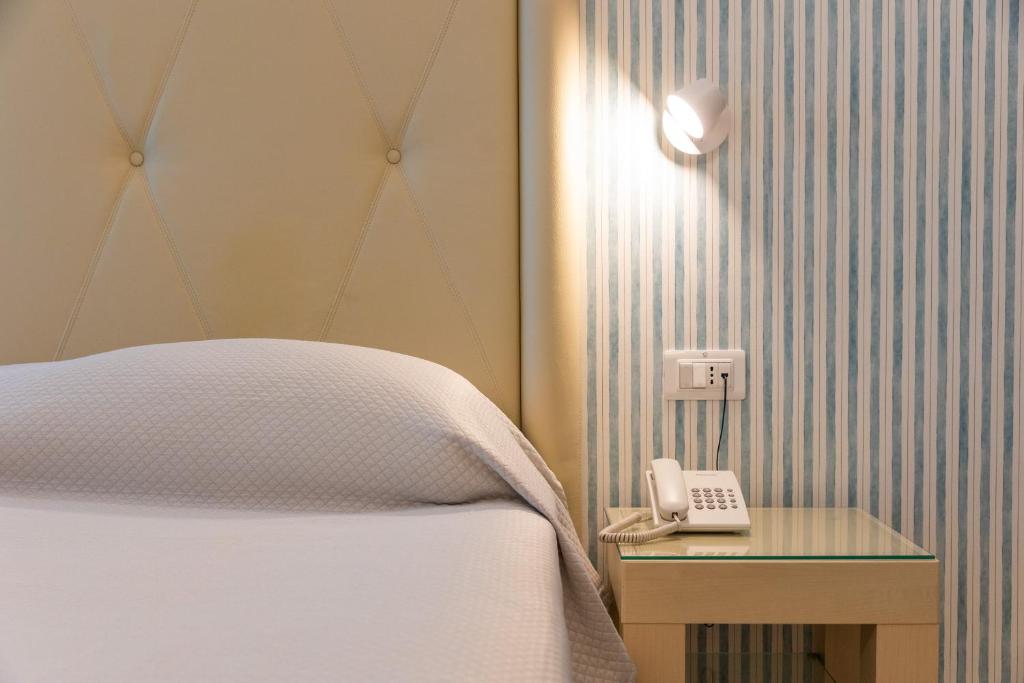 Mūsdienīgs guļamistabas stūris ar mīkstu gultas galvgalu, baltiem palagiem un telefona aparātu uz naktsgaldiņa, fona ar svītrainām tapetēm.