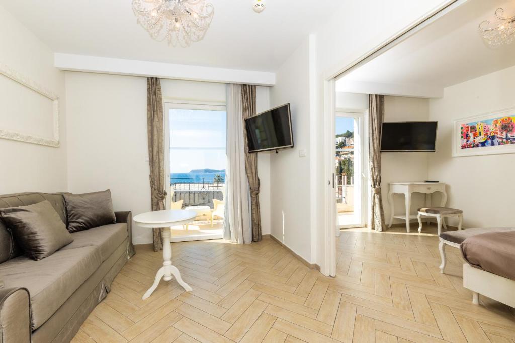 Mūsdienīga viesnīcas istaba ar skatu uz jūru, kurā ir dīvāns, galds un divi televizori. Interjers ir gaišs un elegants, ar koka grīdu un dekoratīvām detaļām.