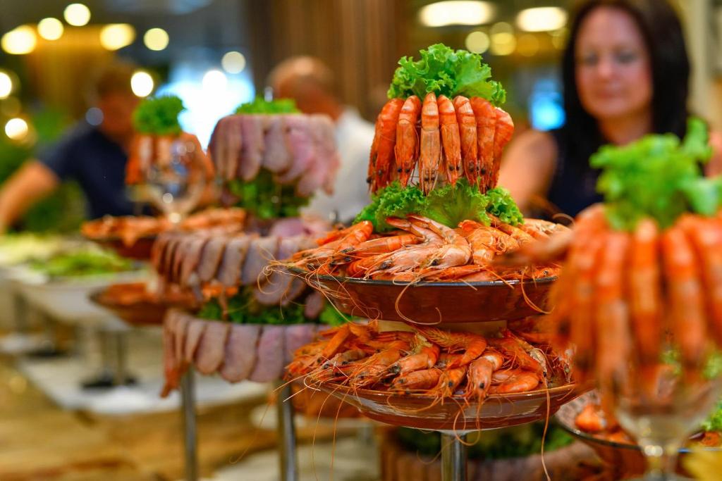 Jūras veltes, tostarp garneles un zivju šķēles, izkārtotas uz vairākām plauktiem, rotātas ar zaļajiem salātiem. Fons ir izsmalcināts restorāns ar gaišu apgaismojumu un ēdotājiem.