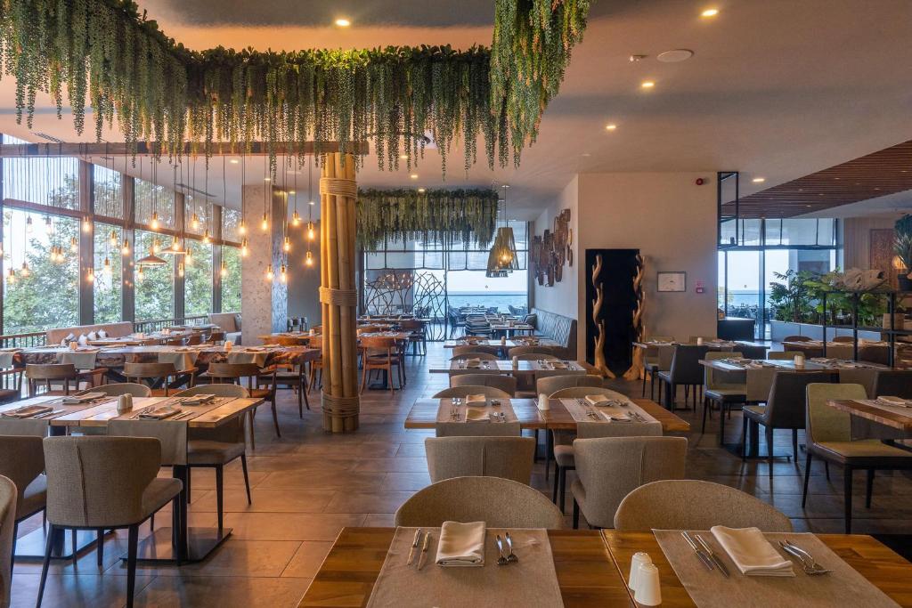 Moderns restorāna interjers ar zaļām augu dekorācijām un dabīgām koka mēbelēm, nodrošinot mājīgu un relaksējošu atmosfēru. Pie logiem skatāma ainava ar dabu un gaismas ķermeņiem, kas piešķir siltumu telpai.