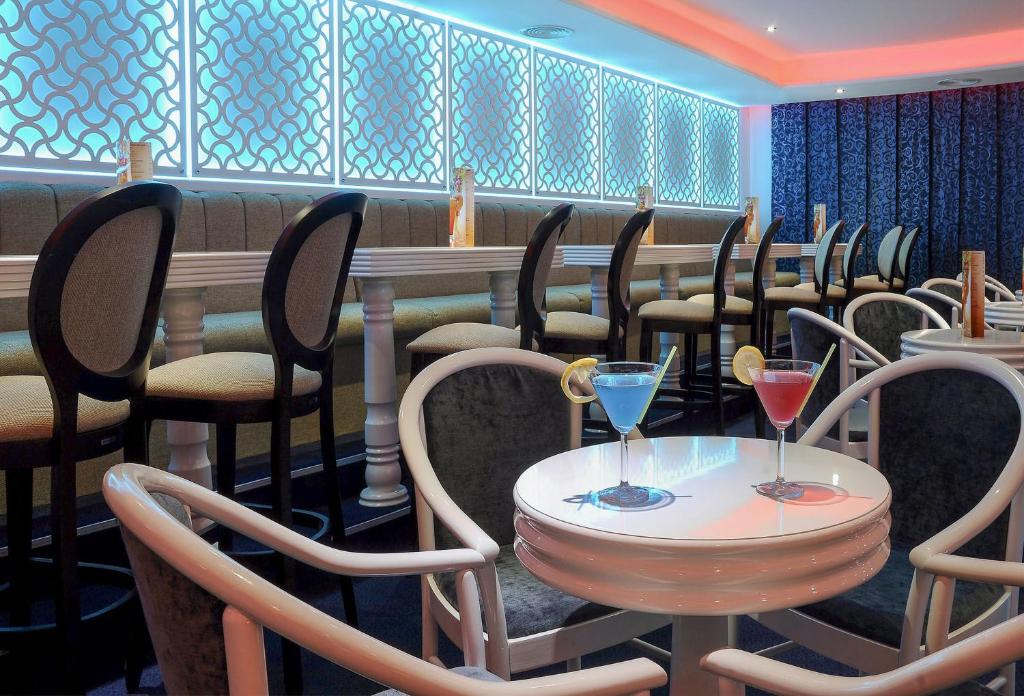 Moderns bārs ar elegantiem krēsliem un divām kokteiļu glāzēm uz galda, kas rada patīkamu atmosfēru. Izsmalcināts interjers ar gaismas elementiem un dekoratīvām sienām.