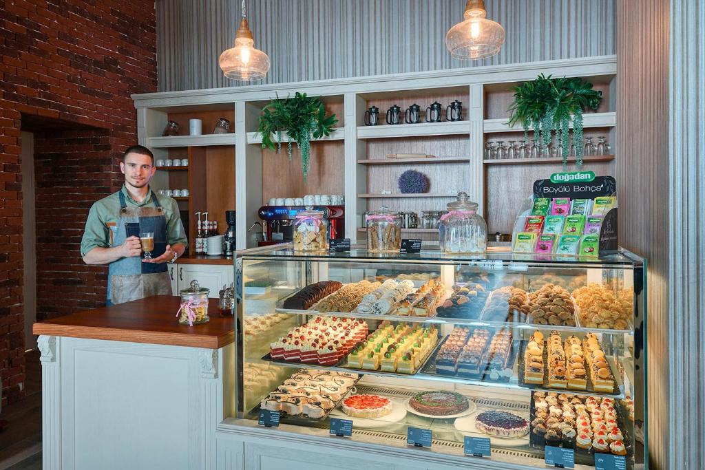 Kafejnīca ar elegantu iekārtojumu un plašu konditorejas izstrādājumu izvēli. Aiz bāra stāv barista, kas pasniedz kafiju un piedāvā gardumus.