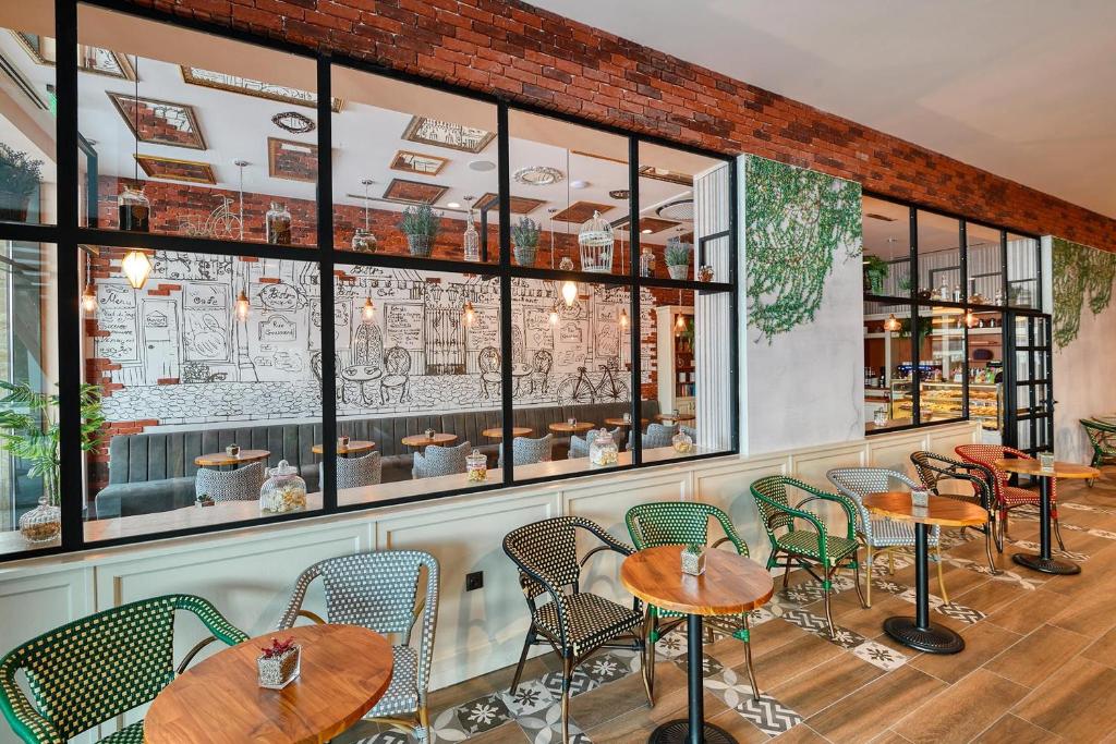 Mūsdienīgs kafejnīcas interjers ar sarkanu ķieģeļu sienu un zīmējumiem uz sienām, kurā redzami koka galdi un krēsli. Svaigas zaļas augu detaļas piešķir dzīvīgumu telpai.