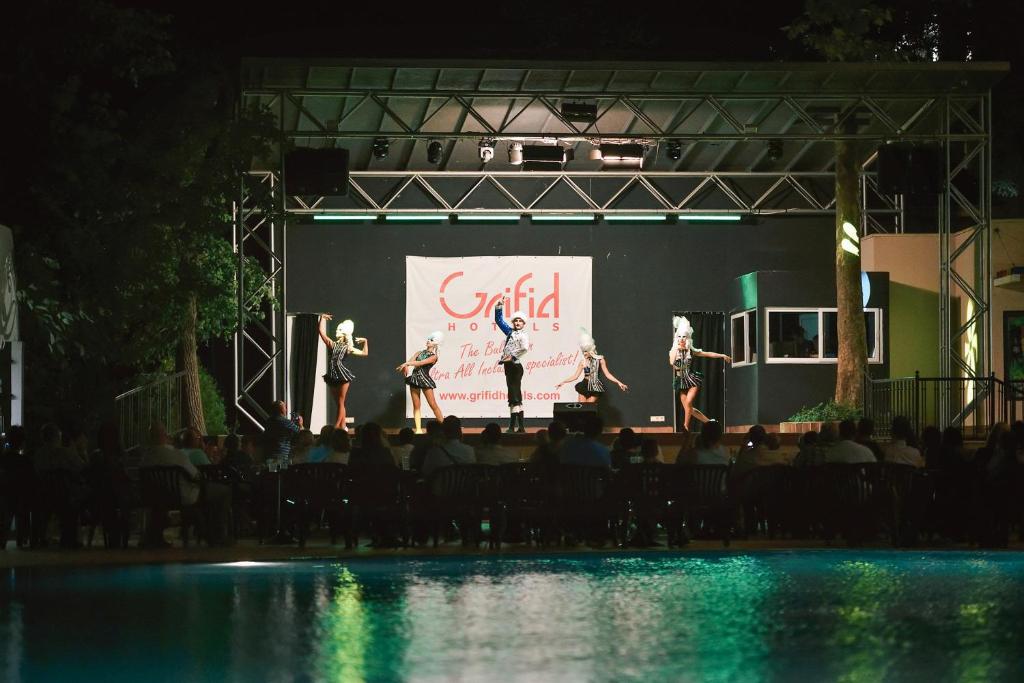 Uz skatuves notiek priekšnesums ar dejotājiem, kuri valkā spilgtas tērpus. Skatītāji bauda izrādi pie baseina. Grifid viesnīcu logo redzams fona plakātā.