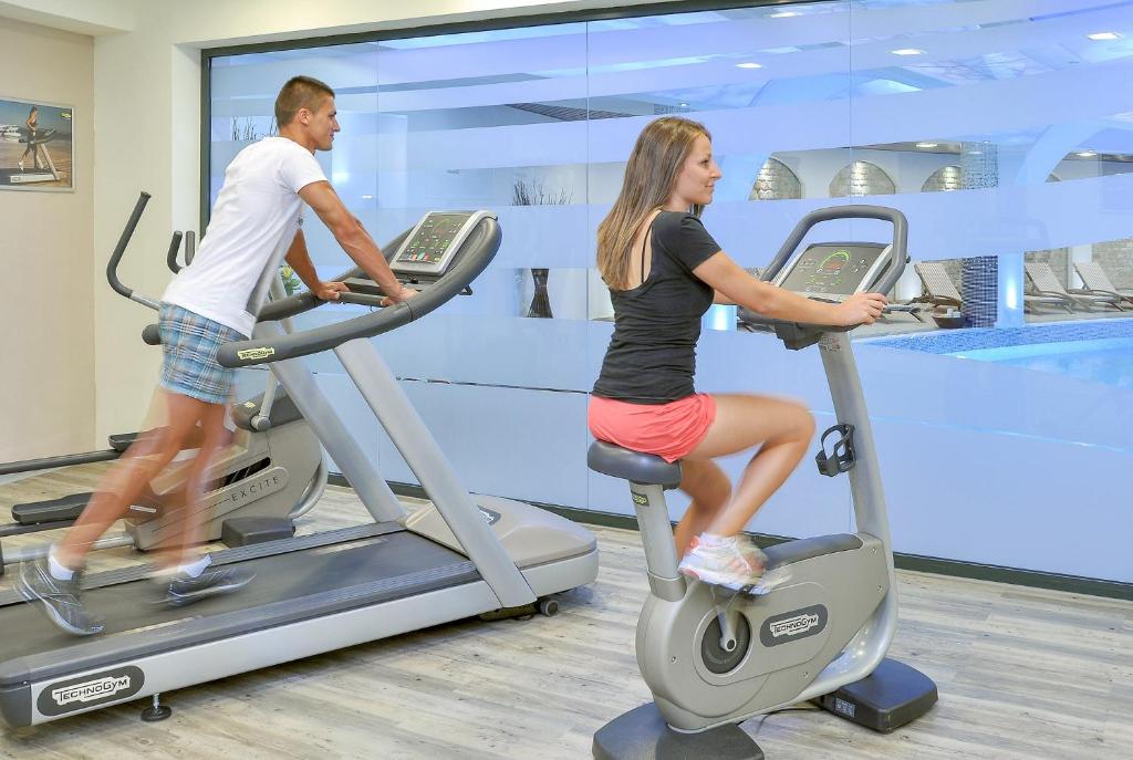 Sieviete un vīrietis vingro sporta zālē, izmantojot dažādus kardio trenažierus, piemēram, elipsveida trenažieri un velotrenažieri, ar skatu uz baseinu.