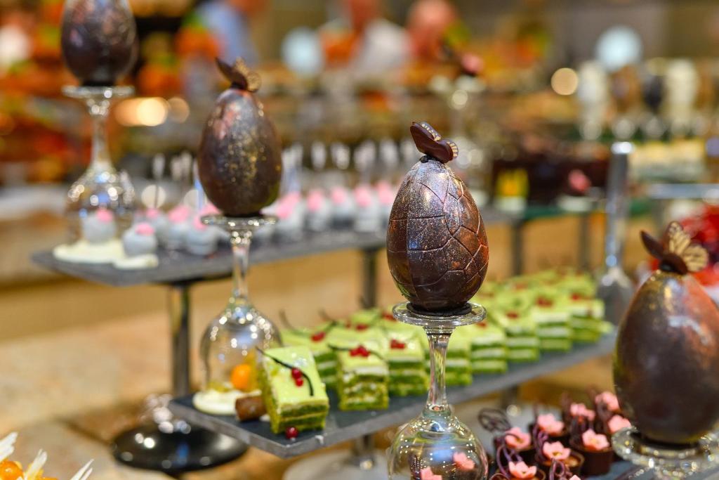 Saldumu displejs ar šokolādes olām un dažādām konditorejas izstrādājumiem, tostarp zaļajiem kūciņām un krēmīgām konfektēm. Eleganti pasniegti uz stendiem, radot krāsainu un pievilcīgu desertu kolekciju.