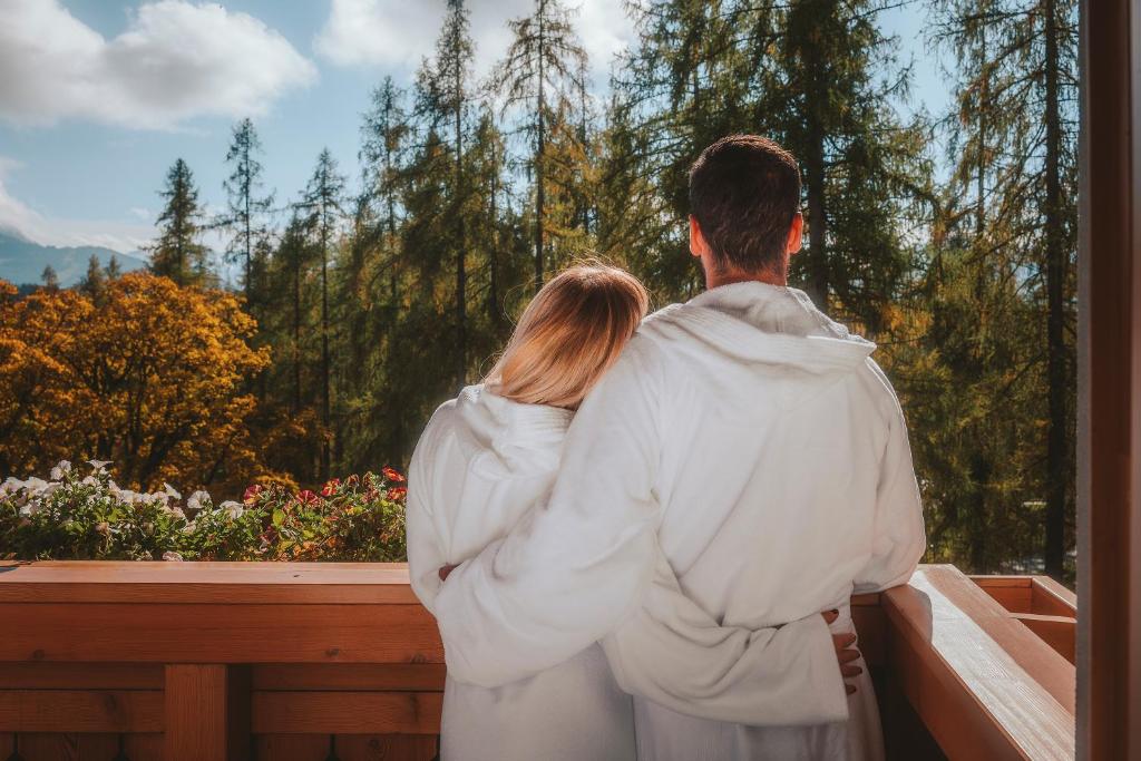 Pāris, tērpies baltos halātos, stāv uz terases un skatās uz ainavisku mežu, apgaismotu saulē, radot romantisku atmosfēru.