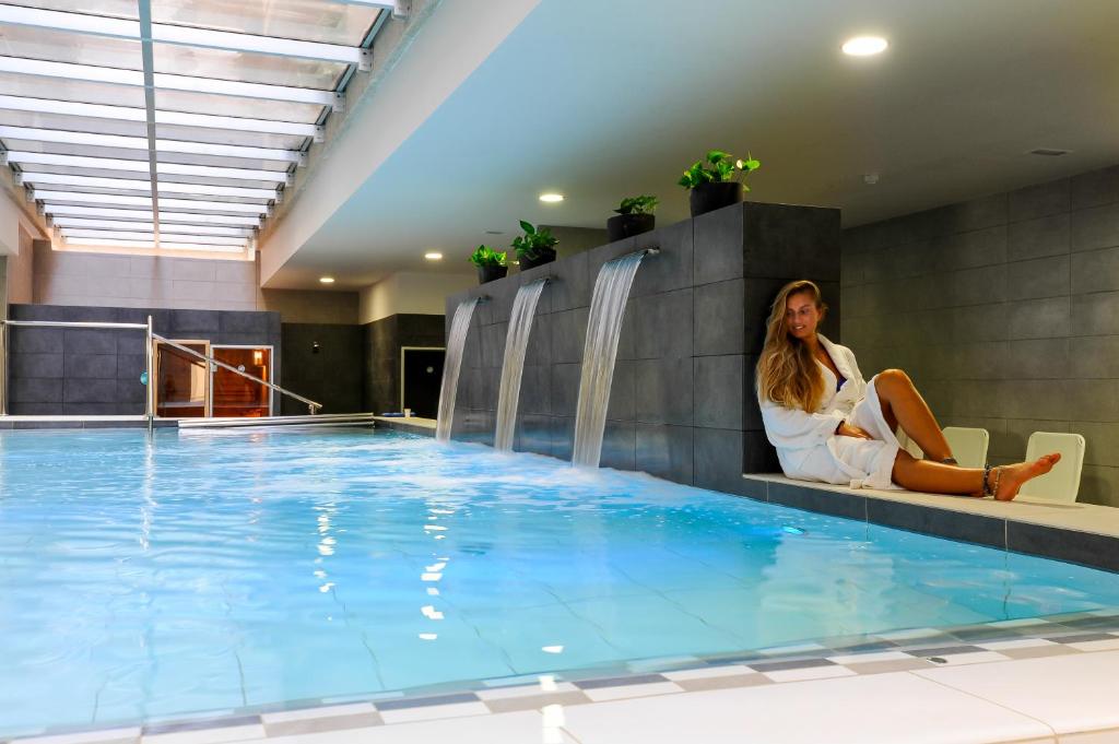 Relaksējoša spa vide ar baseinu un ūdens strūklām, kur sieviete sēž baltā halātā. Gaisma ieplūst caur stikla jumtu, radot mierīgu atmosfēru.