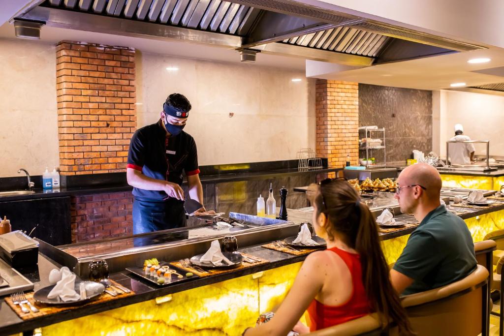 Sushi restorānā šefpavārs gatavo ēdienu pie grila, kamēr apmeklētāji skatās. Restorāna interjers ir moderns, ar siltu apgaismojumu un elegantu dizainu.