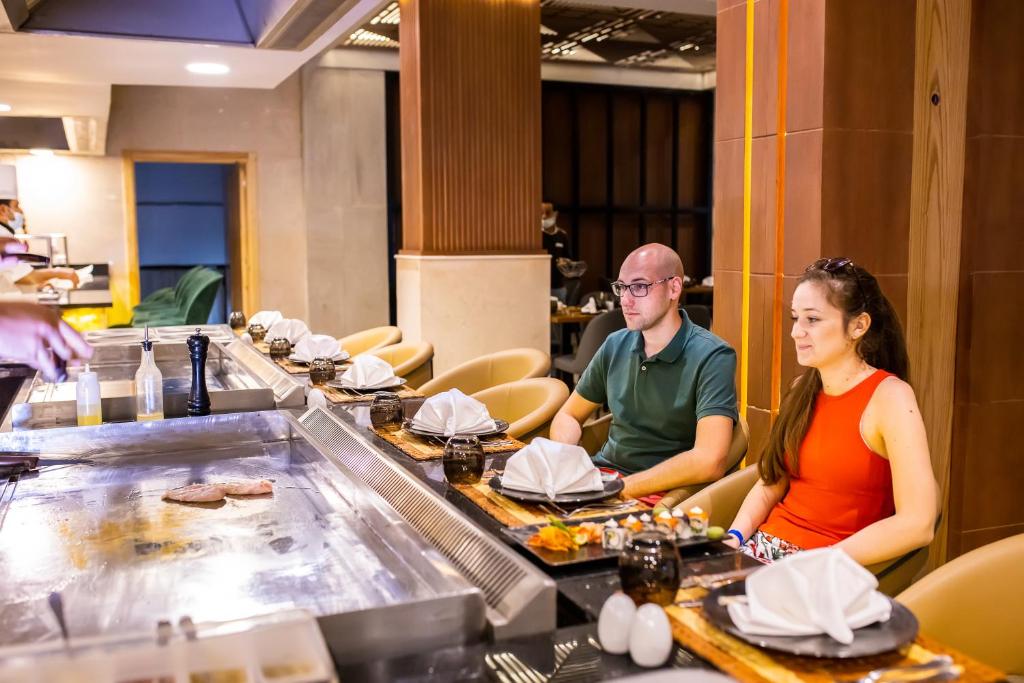 Divi cilvēki sēž pie teppanyaki galda restorānā, gaidot ēdienu, kamēr šefpavārs gatavo maltīti. Uz galda redzami sagatavoti ēdieni un galda piederumi.