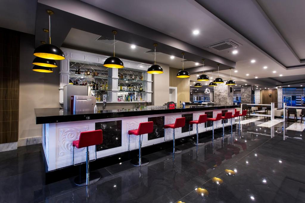 Mūsdienīgs bārs ar melnu marmora virsmu, sarkaniem krēsliem un dzeltenām lampām, kas rada elegantu atmosfēru. Apgaismojums izceļ stikla plauktus ar alkohola dzērieniem.