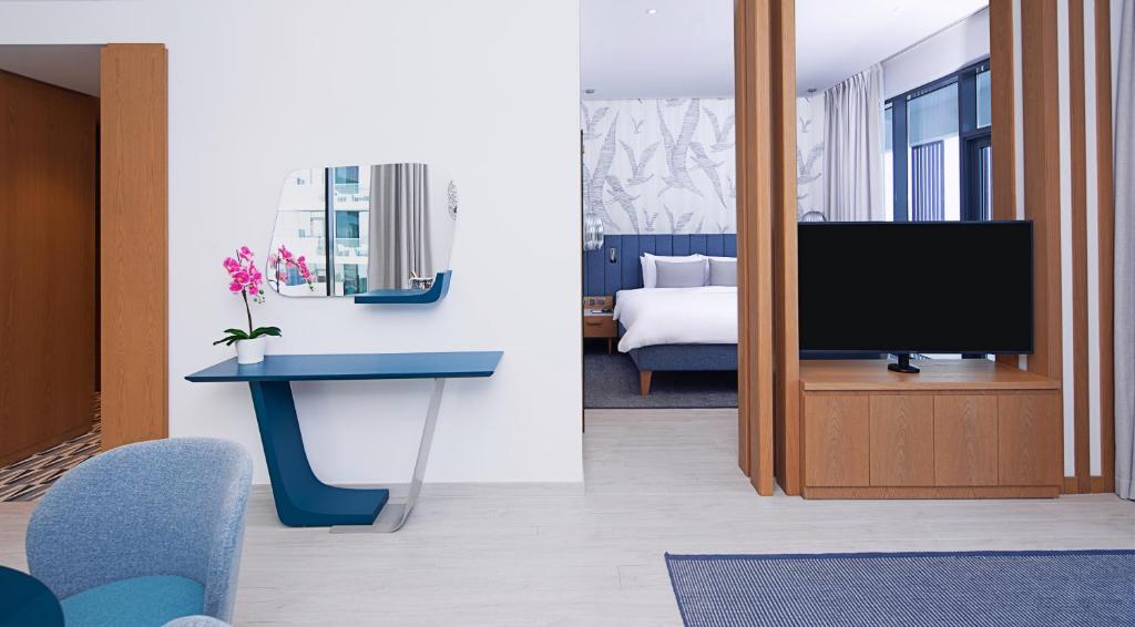 Mūsdienīga viesnīcas istaba ar stilīgu zilu galdu, spoguli un orhideju, redzama arī gulta un televizors fona telpā.