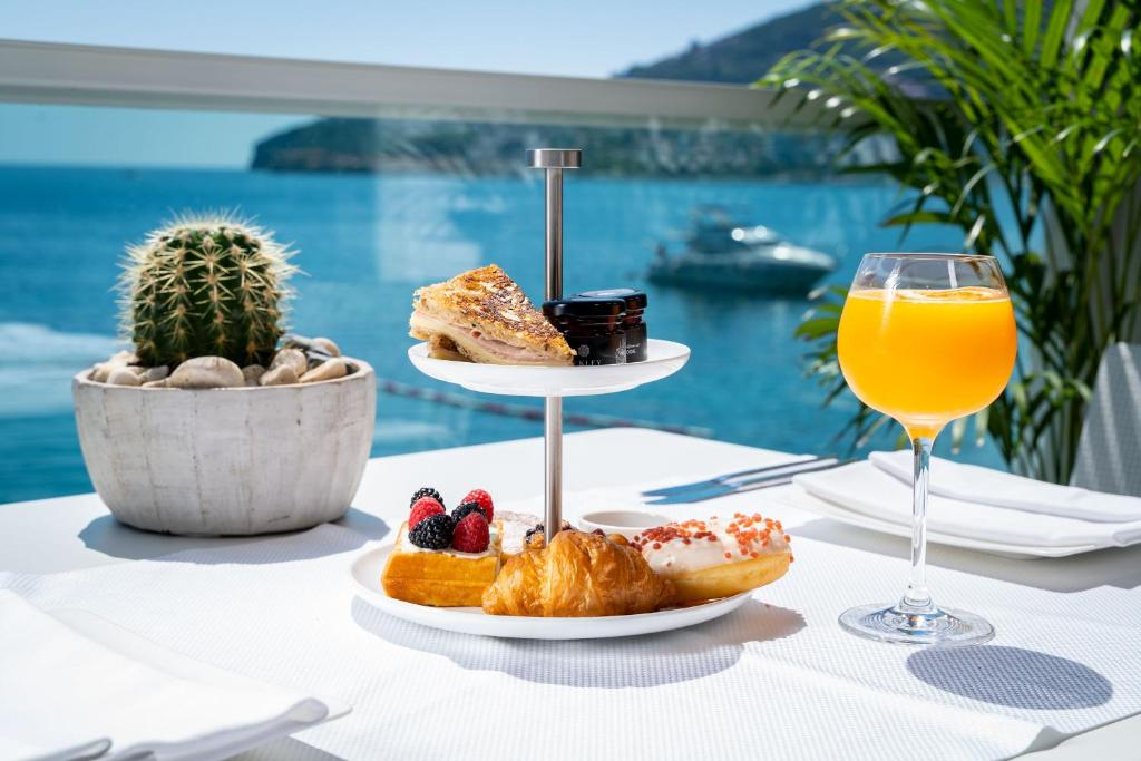 Elegants ēdiens ar desertiem un dzērienu, pasniegts pie galda ar skatu uz jūru. Uz galda ir augļu kokteilis un dažādas kārumi, tostarp kruasāns un saldēti deserti.